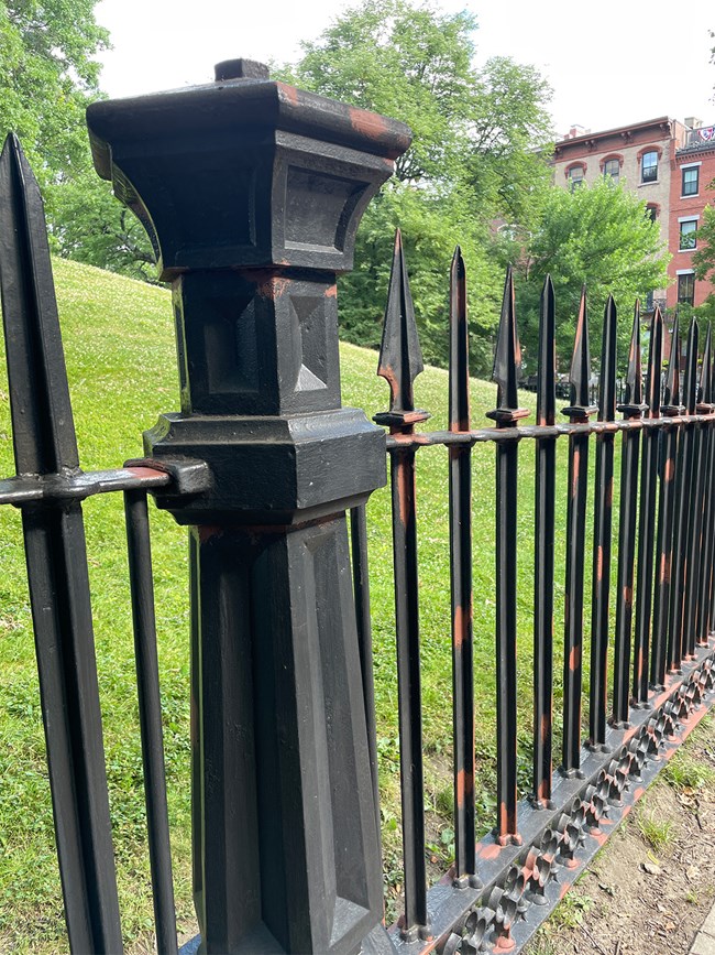 A black wrought-iron fence from a side view. A thick pillar and several smaller bars of the fence are in view, with a floral pattern along the bottom of the fence. The fence has some spots of rust-colored paint applied to it.