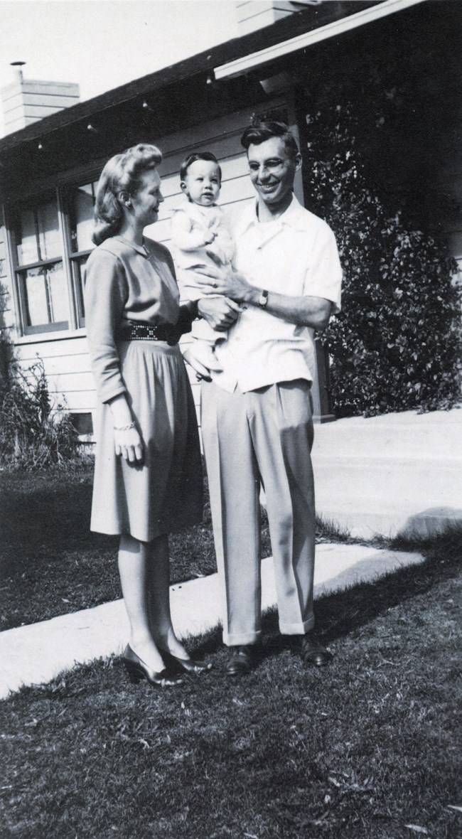 Black and white photo of a young couple with a baby in front of a house