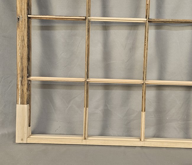 A closeup of the bottom right corner of one of the windows. Most of the wood is dark brown and heavily weathered. The bottom rail, the bottom portion of the left stile, and some of the muntin bars have been replaced by newer, brighter tan wood.