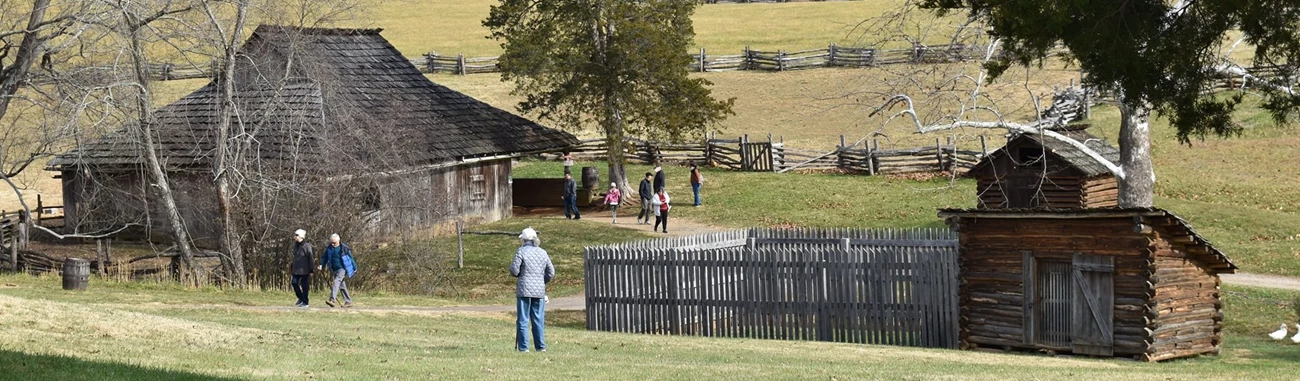 Park visitors walking around a cabin with a split-rail fence in the background.