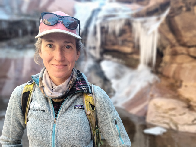 Smiling woman in a baseball cap wearing a backpack and standing in front of a rocky waterfall