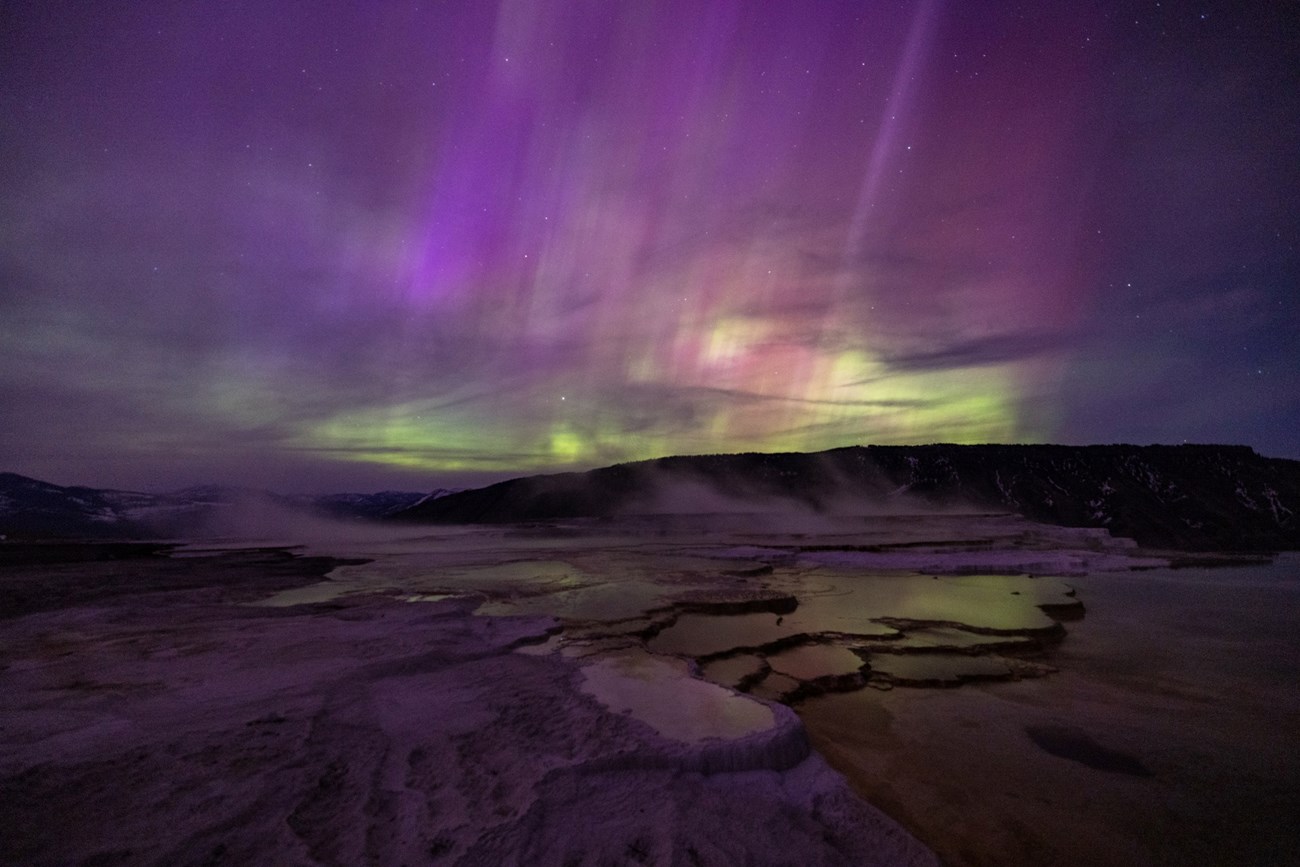 purples, blues, and greens of the northern lights light up the sky over a hydrothermal hot spring at night
