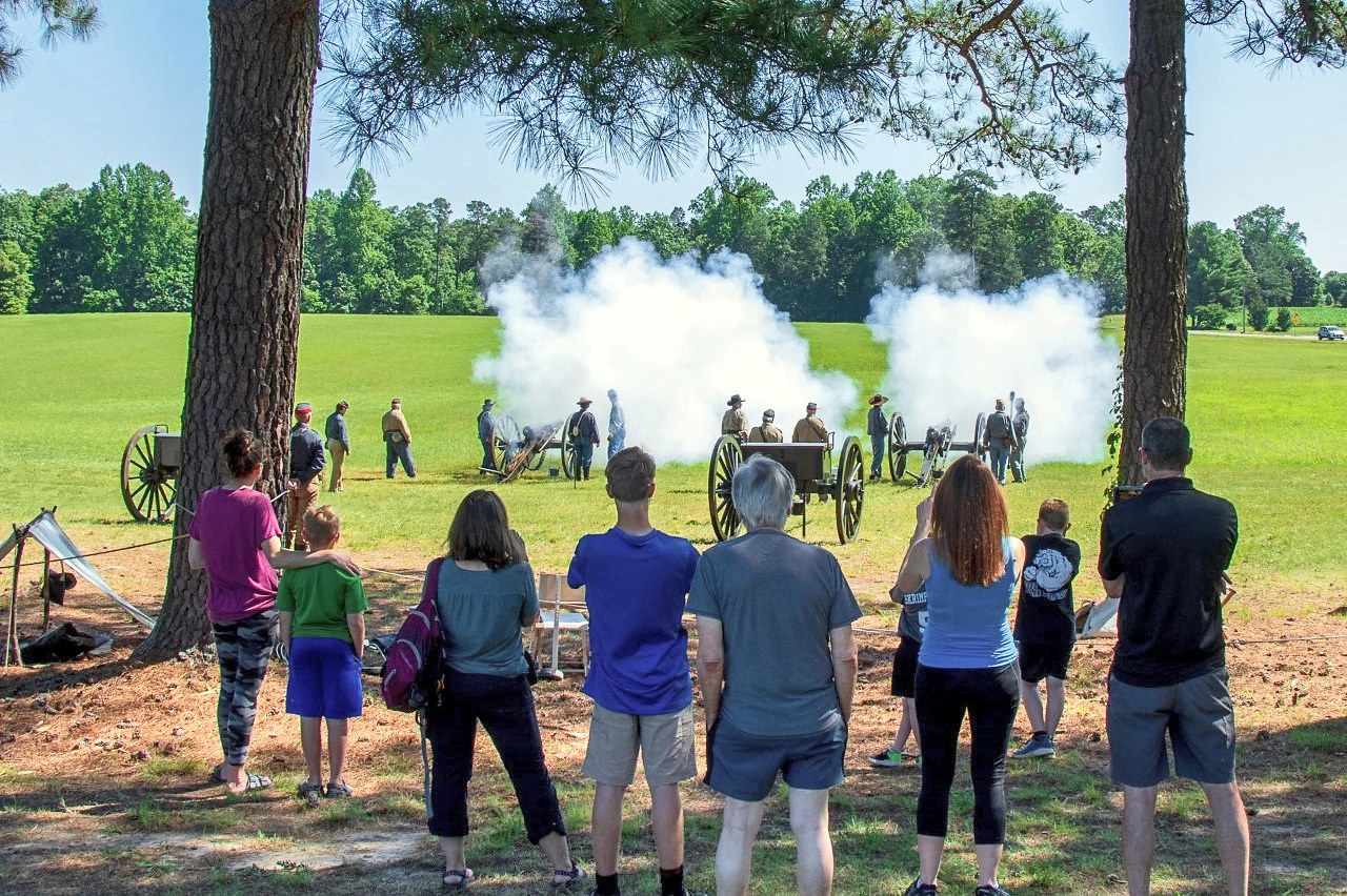 Group of adults and children under a pair of trees, some photographing/filming with mobile devices as clouds of smoke engulf two just-discharged cannons nearby.