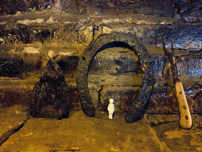 A small ceramic doll stands under a horse shoe and beside an iron head and a fork.