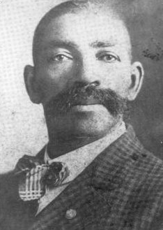 A short haired Black man with a large moustache looking toward the camera with a stoic face, wearing a plaid suit and bow tie.