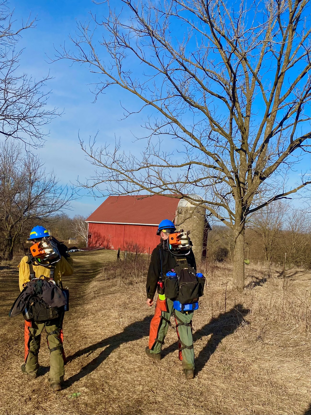 Two people wearing wildland firefighting gear walk along a path with leafless trees and a red barn nearby