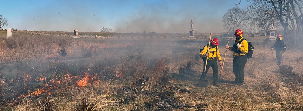 Firefighters use handtools near a fireline with battlefield monuments in the distance.