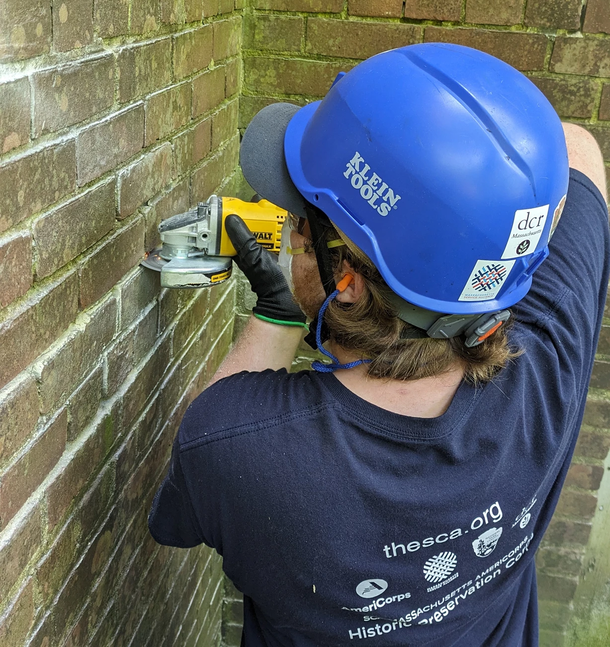 A crew member wearing a hard hat, N95 mask, safety glasses, and earplugs holds an angle grinder up to an old brick wall, removing some of the mortar.