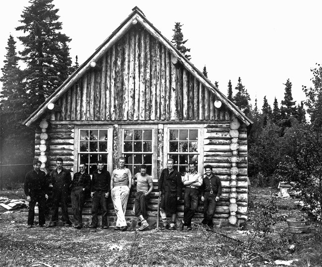 A row of nine men leaning against the front of a log structure