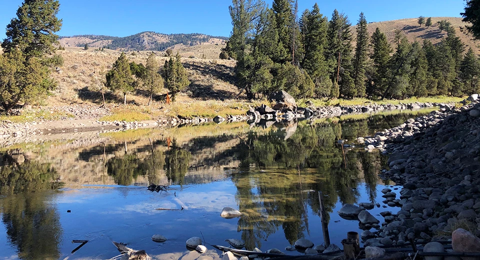 A calm river lined by rocks reflecting the trees and hills behind it.