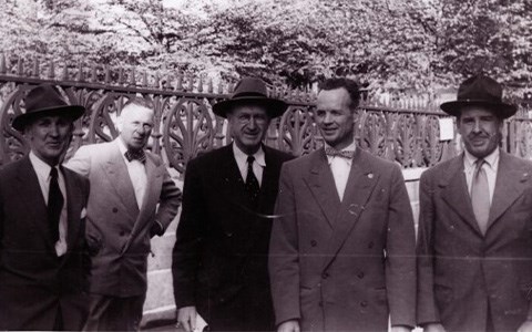 Five men dressed in suits and ties standing in front of an iron fence.