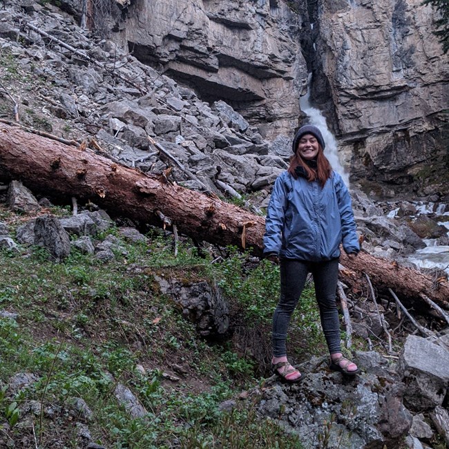 Woman stands on rocky slope with a waterfall streaming from a cliff behind her.