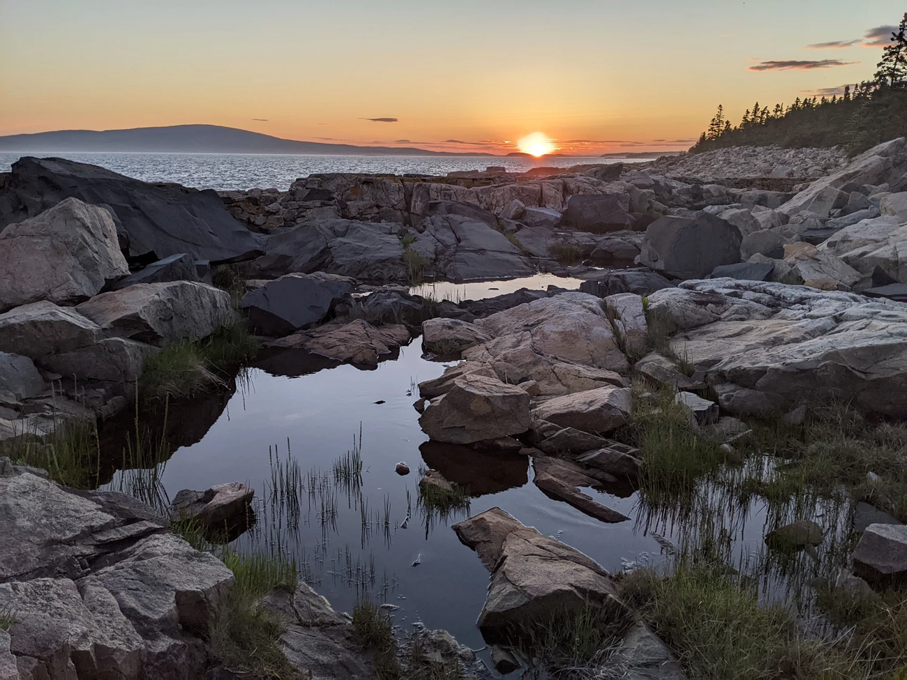A sunset and distant mountain on the horizon, with rocky foreground and pools of calm water reflecting the sky.