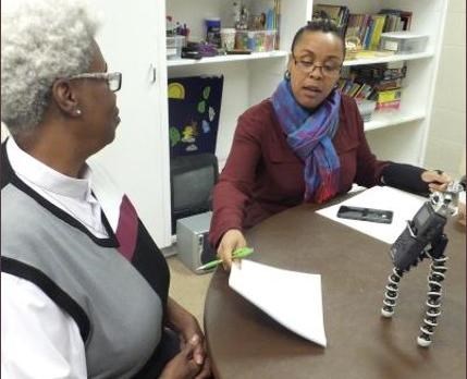 Two people are seated at a table in front of an audio recorder. One woman hands a paper and pen to the other.