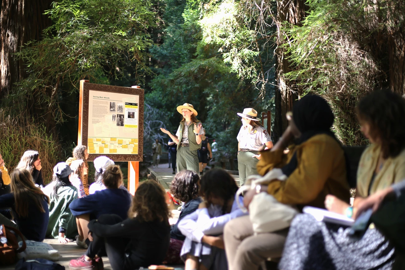 Two rangers in a redwood forest beside a sign, talking with a seated student group