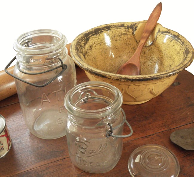 Two glass canning jars and a yellow bowl with a wooden spoon sit at the center of a wooden table. On the left, a can of Underwood Deviled Ham is just visible as is a wooden rolling pin at the top of the photograph.