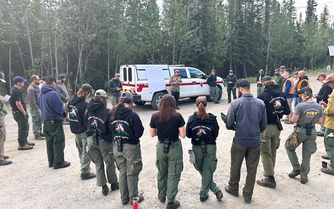 A large group of firefighters gathers for a briefing outside near a fire truck